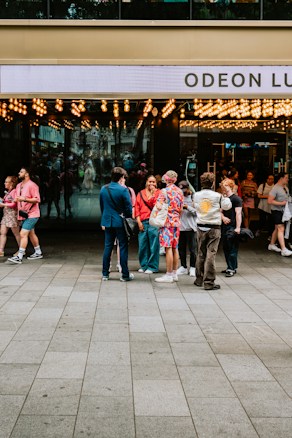 A group of people stand outside a cinema entrance adorned with numerous bright lights. The cinema's name 'Odeon Lux' is partially visible on the sign above the entrance. The individuals appear to be engaged in conversation, with a variety of colorful and casual outfits. The area is busy, with other people visible entering or exiting the venue. The scene suggests a lively, social atmosphere.
