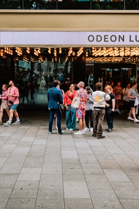 A group of people stand outside a cinema entrance adorned with numerous bright lights. The cinema's name 'Odeon Lux' is partially visible on the sign above the entrance. The individuals appear to be engaged in conversation, with a variety of colorful and casual outfits. The area is busy, with other people visible entering or exiting the venue. The scene suggests a lively, social atmosphere.