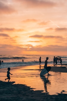Silhouettes of people enjoying the beach at sunset with one person holding a surfboard. The sky is filled with warm hues reflecting on the wet sand, creating a serene and picturesque scene.