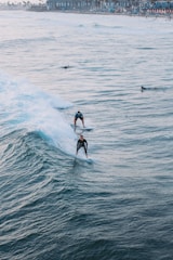 Surfers ride the ocean waves close to a busy beach town. The image captures two surfers skillfully maneuvering through the water, with the coastline visible in the background. The waves are dynamic and create a sense of motion.