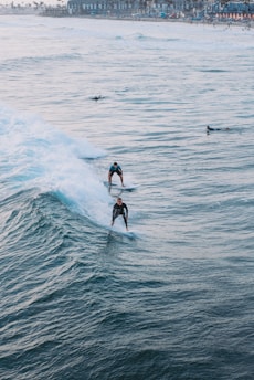 Surfers ride the ocean waves close to a busy beach town. The image captures two surfers skillfully maneuvering through the water, with the coastline visible in the background. The waves are dynamic and create a sense of motion.