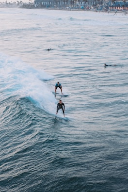 Surfers ride the ocean waves close to a busy beach town. The image captures two surfers skillfully maneuvering through the water, with the coastline visible in the background. The waves are dynamic and create a sense of motion.