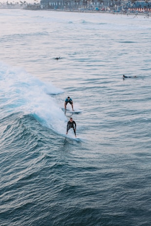 Surfers ride the ocean waves close to a busy beach town. The image captures two surfers skillfully maneuvering through the water, with the coastline visible in the background. The waves are dynamic and create a sense of motion.