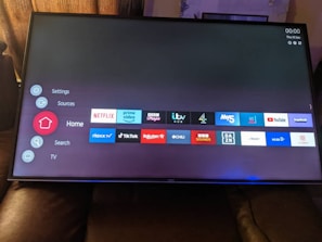 A television screen displays a smart TV interface with multiple app icons arranged in rows. Options such as Settings, Sources, Home, Search, and TV are listed on the left side. Some recognizable app icons include Netflix, Prime Video, BBC iPlayer, ITV Hub, All 4, My5, YouTube, and TikTok. The time and date are shown in the top right corner.