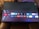 A television screen displays a smart TV interface with multiple app icons arranged in rows. Options such as Settings, Sources, Home, Search, and TV are listed on the left side. Some recognizable app icons include Netflix, Prime Video, BBC iPlayer, ITV Hub, All 4, My5, YouTube, and TikTok. The time and date are shown in the top right corner.