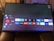 A television screen displays a smart TV interface with multiple app icons arranged in rows. Options such as Settings, Sources, Home, Search, and TV are listed on the left side. Some recognizable app icons include Netflix, Prime Video, BBC iPlayer, ITV Hub, All 4, My5, YouTube, and TikTok. The time and date are shown in the top right corner.