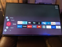 A television screen displays a smart TV interface with multiple app icons arranged in rows. Options such as Settings, Sources, Home, Search, and TV are listed on the left side. Some recognizable app icons include Netflix, Prime Video, BBC iPlayer, ITV Hub, All 4, My5, YouTube, and TikTok. The time and date are shown in the top right corner.
