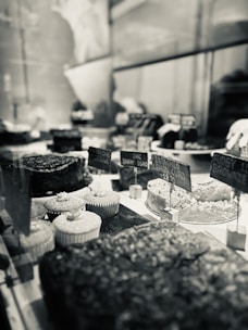 A bakery display with a variety of desserts, including cupcakes, brownies, and cakes. The scene is captured in black and white, emphasizing the textures and details of the treats. Small signs with prices are visible, adding to the rustic yet elegant presentation.