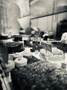 A bakery display with a variety of desserts, including cupcakes, brownies, and cakes. The scene is captured in black and white, emphasizing the textures and details of the treats. Small signs with prices are visible, adding to the rustic yet elegant presentation.