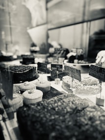 A bakery display with a variety of desserts, including cupcakes, brownies, and cakes. The scene is captured in black and white, emphasizing the textures and details of the treats. Small signs with prices are visible, adding to the rustic yet elegant presentation.