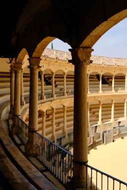 A historic bullring featuring stone columns and arches, with tiered seating and a sandy arena. The architecture displays a classic design with a wooden railing running alongside the pathway, highlighting a sense of symmetry and craftsmanship.