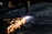 A plasma cutter is in action, emitting bright sparks and a concentrated flame while cutting through a sheet of metal. The metal surface is dark, and the sparks create a striking contrast against it.