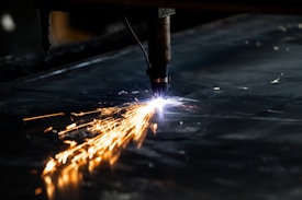 A plasma cutter is in action, emitting bright sparks and a concentrated flame while cutting through a sheet of metal. The metal surface is dark, and the sparks create a striking contrast against it.