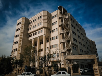 A multi-story hospital building with beige stone or concrete exterior and symmetrical rows of windows. A prominent vertical sign displays the name 'N.H.F HOSPITAL' in large letters. The building also features external staircases and has trees and parked cars in front. The sky is partly cloudy.