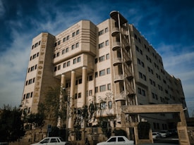 A multi-story hospital building with beige stone or concrete exterior and symmetrical rows of windows. A prominent vertical sign displays the name 'N.H.F HOSPITAL' in large letters. The building also features external staircases and has trees and parked cars in front. The sky is partly cloudy.