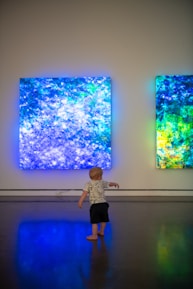 A young child stands on a polished floor, gazing at two large, illuminated abstract art pieces on the wall. The artwork features vibrant shades of blue, green, and hints of yellow, creating a glowing ambiance in an otherwise dimly lit room.