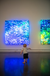 A young child stands on a polished floor, gazing at two large, illuminated abstract art pieces on the wall. The artwork features vibrant shades of blue, green, and hints of yellow, creating a glowing ambiance in an otherwise dimly lit room.