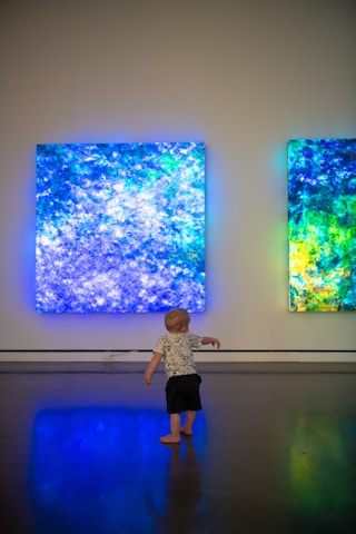 A young child stands on a polished floor, gazing at two large, illuminated abstract art pieces on the wall. The artwork features vibrant shades of blue, green, and hints of yellow, creating a glowing ambiance in an otherwise dimly lit room.
