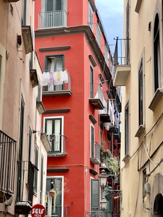 Colorful buildings line a narrow alleyway, with a striking red building prominently in the center. Laundry hangs drying from the balconies, contrasting with the architectural features like shutters and wrought iron railings. A series of lamps and signs are visible along the building walls.