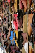 A variety of colorful, handcrafted leather shoes are hanging on display. The shoes come in a range of shades including red, blue, brown, yellow, and black, and feature intricate stitching and punched hole designs. They are neatly arranged in rows, creating a vibrant and artistic pattern.