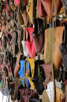 A variety of colorful, handcrafted leather shoes are hanging on display. The shoes come in a range of shades including red, blue, brown, yellow, and black, and feature intricate stitching and punched hole designs. They are neatly arranged in rows, creating a vibrant and artistic pattern.