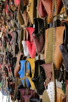 A variety of colorful, handcrafted leather shoes are hanging on display. The shoes come in a range of shades including red, blue, brown, yellow, and black, and feature intricate stitching and punched hole designs. They are neatly arranged in rows, creating a vibrant and artistic pattern.