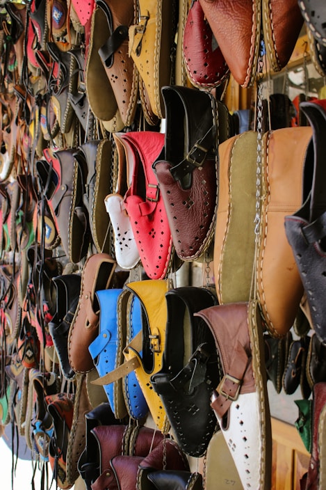 A variety of colorful, handcrafted leather shoes are hanging on display. The shoes come in a range of shades including red, blue, brown, yellow, and black, and feature intricate stitching and punched hole designs. They are neatly arranged in rows, creating a vibrant and artistic pattern.