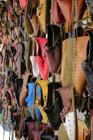 A variety of colorful, handcrafted leather shoes are hanging on display. The shoes come in a range of shades including red, blue, brown, yellow, and black, and feature intricate stitching and punched hole designs. They are neatly arranged in rows, creating a vibrant and artistic pattern.