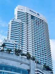 A tall modern hotel building with numerous windows and a sleek architectural design. The hotel name is visible at the top, and there are palm trees adorning the lower levels. The sky is clear with a few white clouds.