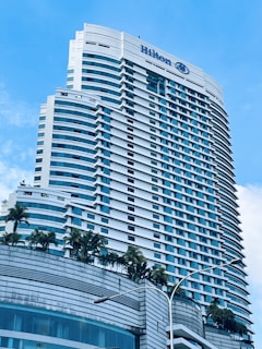 A tall modern hotel building with numerous windows and a sleek architectural design. The hotel name is visible at the top, and there are palm trees adorning the lower levels. The sky is clear with a few white clouds.