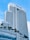A tall modern hotel building with numerous windows and a sleek architectural design. The hotel name is visible at the top, and there are palm trees adorning the lower levels. The sky is clear with a few white clouds.