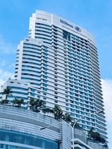 A tall modern hotel building with numerous windows and a sleek architectural design. The hotel name is visible at the top, and there are palm trees adorning the lower levels. The sky is clear with a few white clouds.