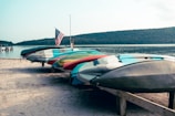 Several kayaks are neatly lined up on a sandy beach, with an American flag standing tall beside them. The background features a calm lake surrounded by tree-covered hills under a clear sky.