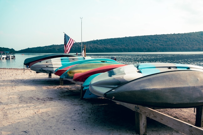 Several kayaks are neatly lined up on a sandy beach, with an American flag standing tall beside them. The background features a calm lake surrounded by tree-covered hills under a clear sky.