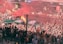 A large crowd of enthusiastic fans fills the stands, with many people dressed in red clothing and waving flags. There is a vibrant display of smoke flares emitting red smoke above the crowd. The atmosphere appears lively and energetic, enhanced by spectators raising their hands in excitement. A fence separates the spectators from an area occupied by event staff.