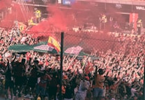 A large crowd of enthusiastic fans fills the stands, with many people dressed in red clothing and waving flags. There is a vibrant display of smoke flares emitting red smoke above the crowd. The atmosphere appears lively and energetic, enhanced by spectators raising their hands in excitement. A fence separates the spectators from an area occupied by event staff.