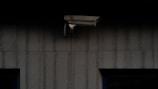 A security camera is mounted on a dark, textured wall. The camera is positioned to monitor an area, possibly outdoor or in a low-light environment.