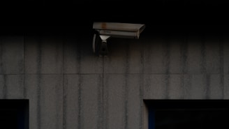 A security camera is mounted on a dark, textured wall. The camera is positioned to monitor an area, possibly outdoor or in a low-light environment.