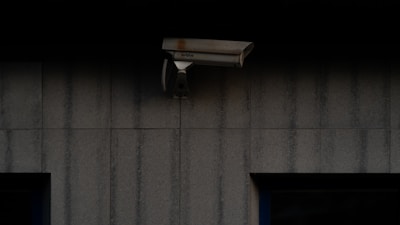 A security camera is mounted on a dark, textured wall. The camera is positioned to monitor an area, possibly outdoor or in a low-light environment.