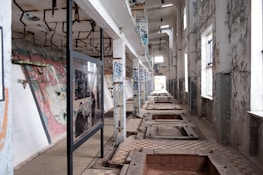 An abandoned industrial building with peeling paint and graffiti on the walls. The room features large windows on one side, allowing natural light to illuminate the decay and urban artwork. There are empty pits on the floor and a series of rusted metal beams suspended from the ceiling.