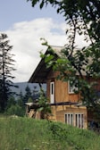 A wooden house is nestled in a lush green landscape with tall trees in the background. The sky is partly cloudy, and the foreground features branches with green leaves, creating a serene and natural setting.