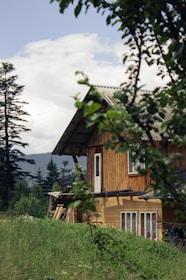 A wooden house is nestled in a lush green landscape with tall trees in the background. The sky is partly cloudy, and the foreground features branches with green leaves, creating a serene and natural setting.