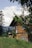 A wooden house is nestled in a lush green landscape with tall trees in the background. The sky is partly cloudy, and the foreground features branches with green leaves, creating a serene and natural setting.
