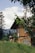 A wooden house is nestled in a lush green landscape with tall trees in the background. The sky is partly cloudy, and the foreground features branches with green leaves, creating a serene and natural setting.