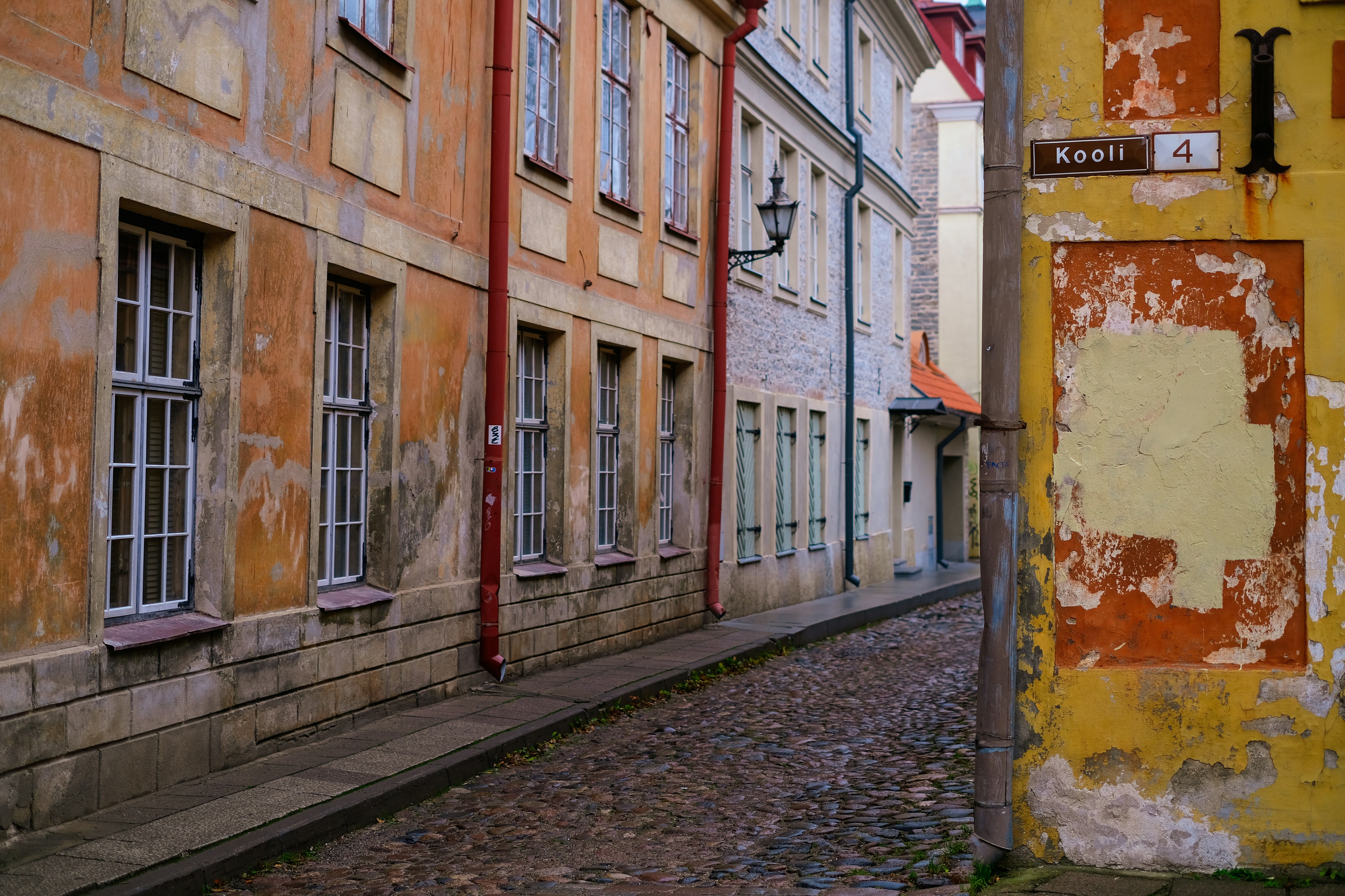 a narrow street with old buildings on both sides, A charming cobblestone street in Tallinn, Estonia, specifically in the historic Old Town. The buildings lining the street are characterized by their weathered facades, with shades of warm orange and yellow, and feature traditional architectural details such as large windows and decorative trim. A street sign indicates the location as Kooli 4.