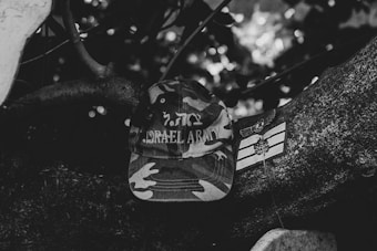 A camouflage-patterned cap with the words 'Israel Army' embroidered on it is placed on a tree branch. Next to the cap is a patch with military symbols. The background is filled with foliage and blurred natural elements.