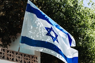 A flag with blue and white colors is displayed outdoors, prominently featuring a blue Star of David in the center. The flag is waving slightly in front of a background filled with lush green leaves and branches. The scene includes a structure with a decorative wall, adding architectural interest.