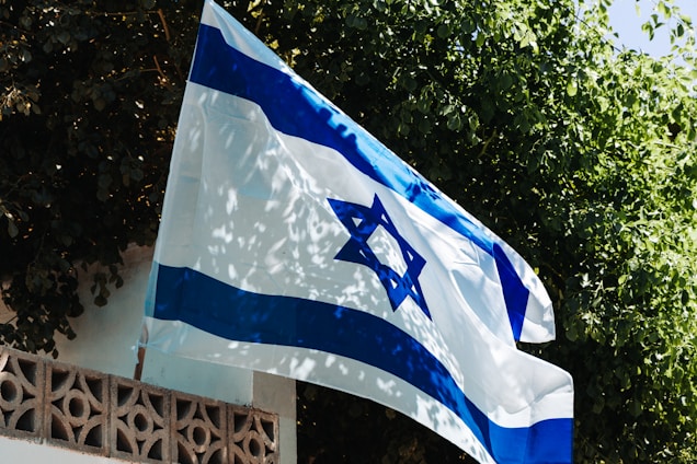 A flag with blue and white colors is displayed outdoors, prominently featuring a blue Star of David in the center. The flag is waving slightly in front of a background filled with lush green leaves and branches. The scene includes a structure with a decorative wall, adding architectural interest.