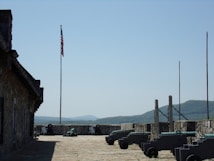 A historic fort setting with a row of cannons lined up against a stone wall. An American flag is flying on a tall flagpole, with mountains visible in the distance under a clear blue sky. The setting suggests a military atmosphere with an emphasis on history and heritage.