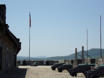 A historic fort setting with a row of cannons lined up against a stone wall. An American flag is flying on a tall flagpole, with mountains visible in the distance under a clear blue sky. The setting suggests a military atmosphere with an emphasis on history and heritage.